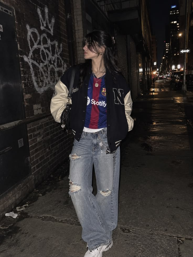 Streetwear blokecore outfit featuring an FC Barcelona football jersey layered under a vintage varsity jacket and ripped jeans.