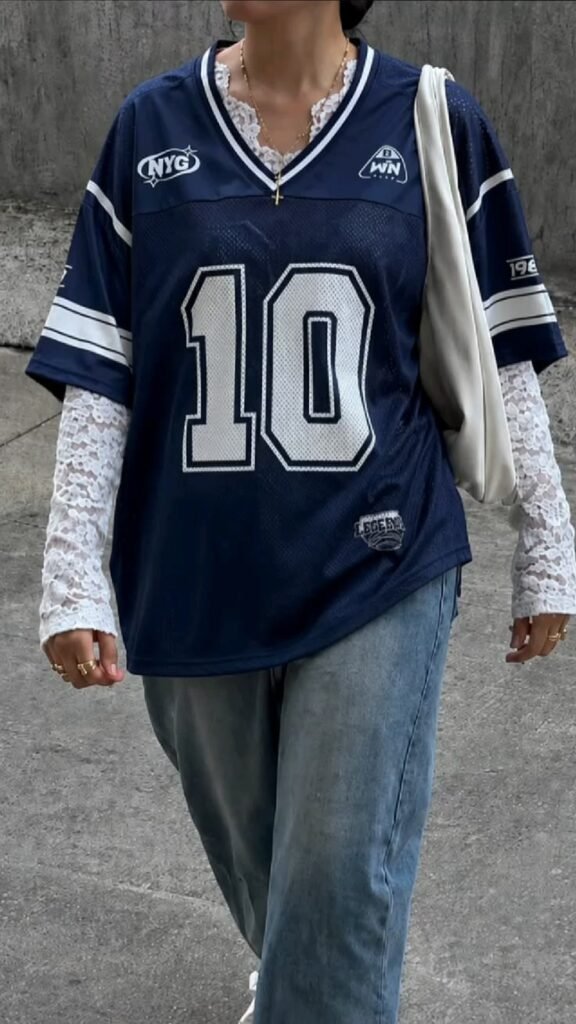 Streetwear blokecore aesthetic featuring a dark blue jersey layered over a white lace long-sleeve top with relaxed denim jeans.