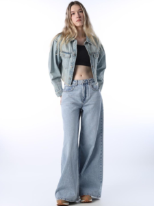 Full-length view of a woman posing against a white studio background wearing a light-wash denim cropped jacket layered over a black crop top, styled with high-waisted, wide-leg light blue jeans.