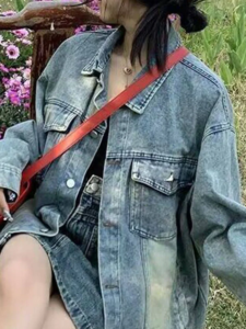 Cropped portrait shot of a woman outdoors surrounded by pink flowers, wearing a light blue denim jacket styled with a thin gold chain necklace and a bright red bag strap slung across her chest.
